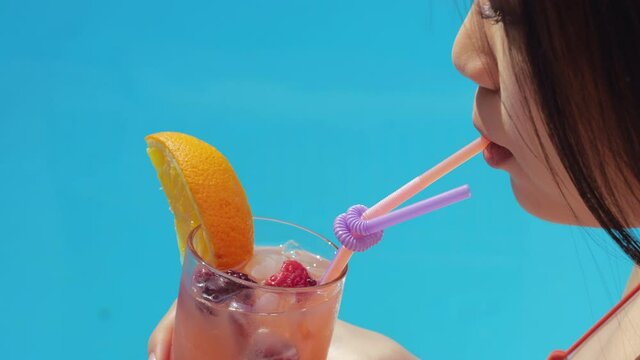 Side view of woman hand body holding glass with ice fresh orange lemonade and joyful pretty young face. Close up Asia female lips drinking non-alcoholic fruit cocktail through straw near pool