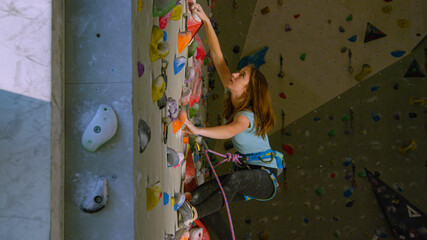 CLOSE UP: Nimble girl loops rope in a carabiner while climbing up an indoor wall