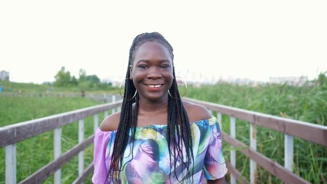 attractive smiling African-American girl with cornrows and large golden earrings in colorful dress walks along wooden bridge among fields