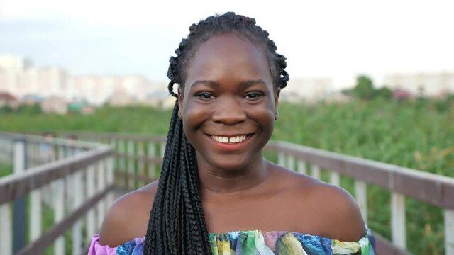 attractive African-American girl with long cornrows in bright color blouse smiles standing on wooden bridge in green reeds closeup slow motion