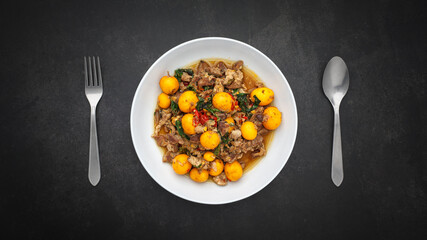 top view of hot and spicy stir fried chicken ovary egg with chicken entrails and basil beside spoon and fork on dark grey texture background with copy space, Thai local street food