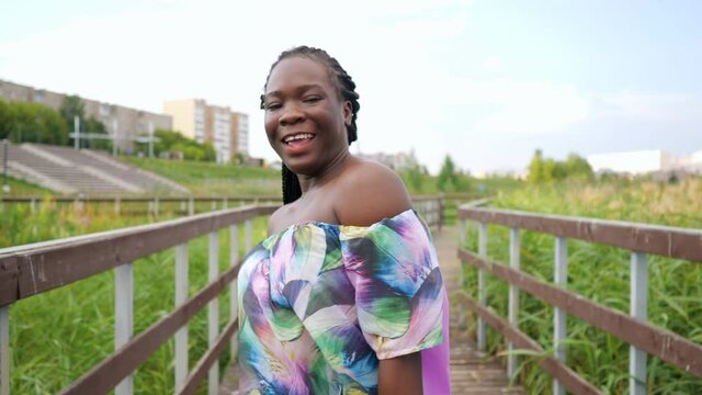 positive active African-American woman with cornrows in elegant blouse dances walking along empty wooden bridge among park reeds slow motion