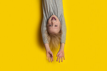 Surprised fair-haired boy hanging upside down with arms outstretched. Portrait of child on bright...