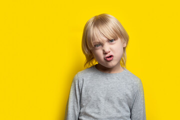 Funny boy with long blond hair poses faces. Portrait of child on yellow background