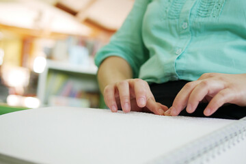 Close-up of blind person woman hands reading Braille book studying in creative library. Braille is...