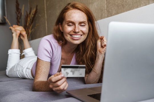 Image of redhead cheerful woman holding credit card and using laptop