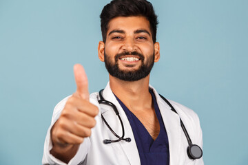 young Indian / Asian doctor with stethoscope in white coat showing thumb up on blue background.