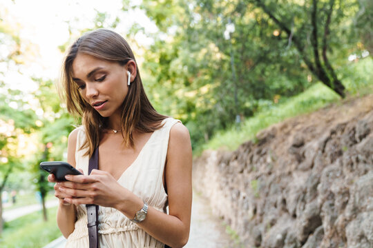 Image of young woman wearing earbuds holding cellphone in park