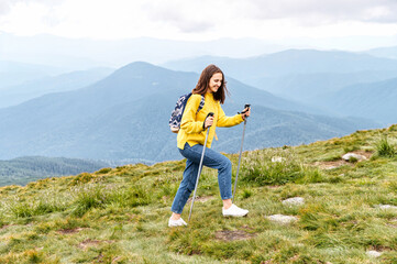 Naklejka na ściany i meble A cheerful hiker girl in yellow sweater with a backpack walks with a sticks on mountains footpath with a scenic view on the background