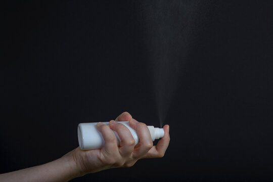 Female hand holding white sprey bottle and spray on dark background 