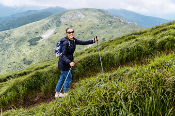 A cheerful hiker girl with a backpack walks with a sticks on mountains footpath with a scenic view...