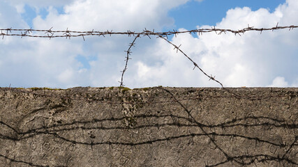 Old rusty barbed wire on an old concrete wall, bright sunny day, blue sky with white clouds in the background. Symbol of lost freedom