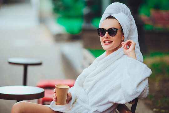 A girl in a robe and a towel on her head sits drinking coffee