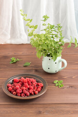 Red ripe fresh raspberries in a brown plate and a bunch of Melissa in a mug on the wooden table