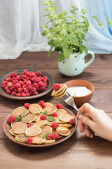Pancake cereals in your hand on a fork with a berry. Red ripe fresh raspberries in a brown plate and a bunch of Melissa in a mug on the dark wooden table near the window. Delicious Breakfast