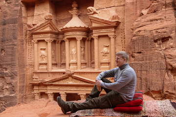Male caucasian tourist sits on the cliff during winter day. Temple Al-Khazneh (The Treasury) carved...