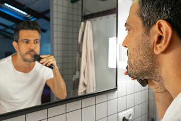 handsome man is shaving his beard with trimmer machine in front of bathroom mirror