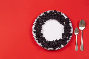 Black and blue ripe blackberries, laid out in a circle on a white plate, in the center of an empty space, two spoons on a red background. Healthy dessert. Space for text. Top view. Mockup