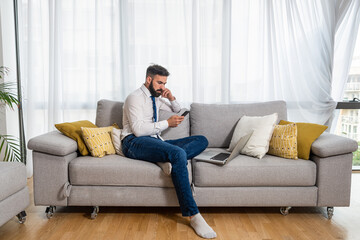 Obraz premium Young freelancer business man sitting on sofa at his home and taking a break on his phone of laptop and waiting for his boss to talk about new business project as freelance worker 