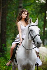 Young beautiful girl with white horse at summer green forest. Caucasian woman horseback rider in dress in boho style. Summertime scene