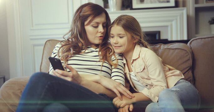 Cheerful Mom With Small Cute Kid Daughter Looking At Screen Phone. Woman Holding Smartphone In Hands And Talking With Child Sitting On Couch.