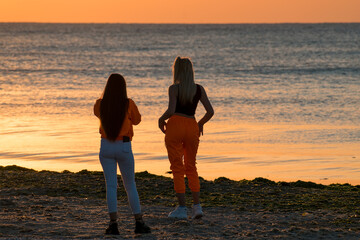 Two young women on the beach, at sunrise, take pictures of each other. Rear view.