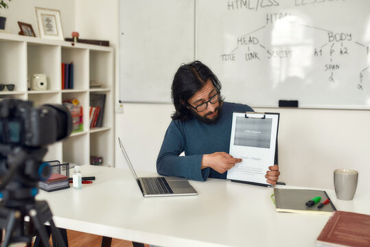 Young caucasian man teacher wearing eyeglasses teaching how to code HTML, sitting at his workplace and giving online lesson. Recording video blog