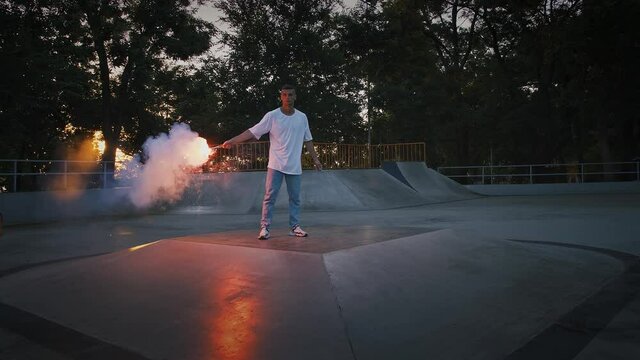 Young man in casual clothes is holding glowing red signal flare and going to perform breakdance on pump track of skatepark. Slow motion