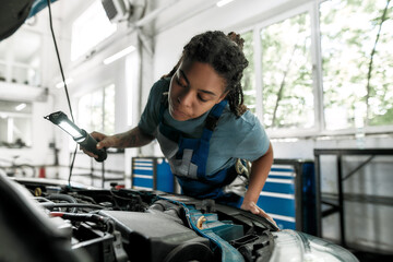 Moments of caring. Young african american woman, professional female mechanic looking, examining under hood of car with torch at auto repair shop