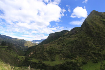 A hilly or mountainous landscape with the several hills covered in grass and small bushes and a bright blue sky with a few white clouds