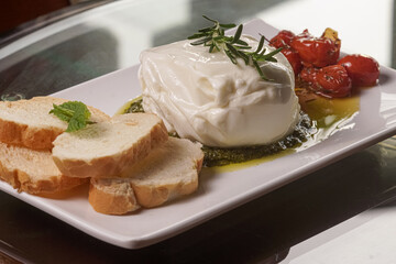 Burrata ornamented with a rosemary branch, presented with olive oil, pesto sauce, toast and tomatoes in a white tray on a glass top. Close up photo.