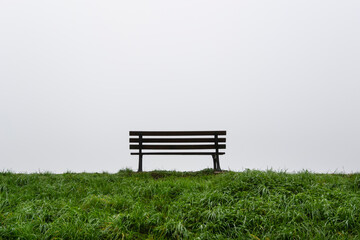 An empty lonely bench on green grass with grey, foggy sky in the background. Copy space.