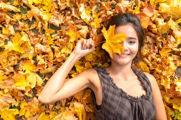 Young happy Asian woman smiling while covering face with leaf lying down on yellow autumn leaves