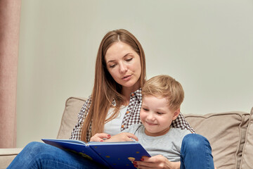 Young mom and son are reading a book together while sitting at home on the couch