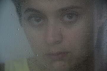 Portrait of a young brunette woman behind glass with rain drops. The concept of depression, blurring about the future, determination. Focus on drops on glass.