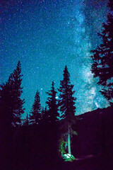 The Milky Way galaxy shines in the night sky over a small illuminated camping tent nestled among the trees.