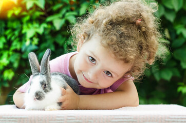 little beautiful girl smiling, hugging a baby rabbit on the green background in summer. happy laughing child and pet playing outdoors. female looking in the camera. bunny is a symbol of Easter.