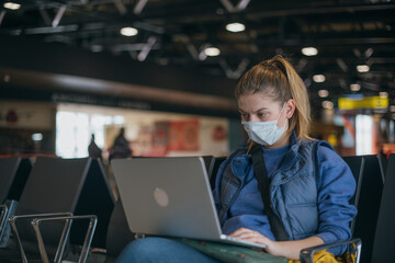 A woman is sitting at the airport with a laptop in a medical mask