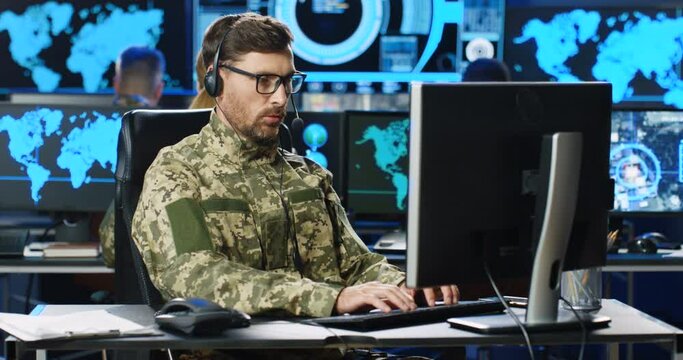 Caucasian male army officer in glasses and headset talking and working on computer in analytic secret base in military. Man militarian official typing in dark office. Digital support. Surveillance.