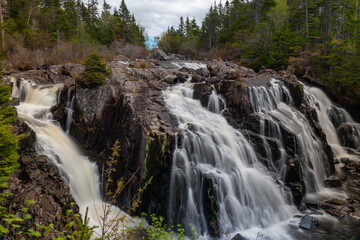 Multiple long natural waterfalls over a dark brown jagged rock cliff. The water is soft and white with some brown silt flowing over the edges. The background is blue sky, clouds and coniferous trees.