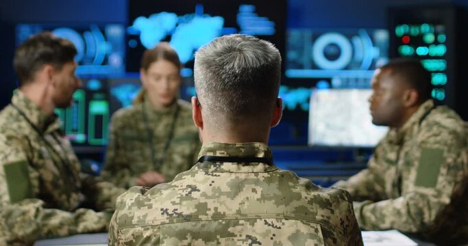 Rear of mixed-races militarian council sitting at table in uniforms, deciding war or defense tactics in dark monitoring room. Back view of multiethnic people, officers talking, discussing terrorism.