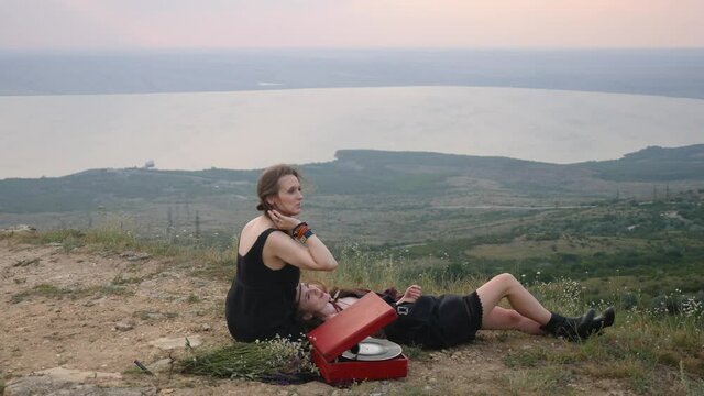 two hipster girls lie on the ground in the mountains and listen to music from a gramophone