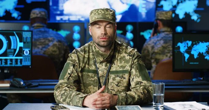 Portrait of Caucasian handsome man military spokesman in uniform sitting in front of camera and telling last war news in monitoring center. Screens on background. Male public representative of troops.