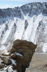 Some stones have been piled beside a mountain slope which is covered in snow. Patches of snow are also on the rocks. The surrounding landscape is rocky. The sky is blue.