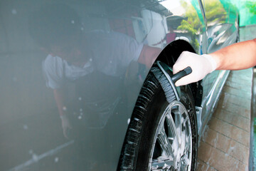 Asian worker in car care garage cleaning a dirty vehicle wheels by using brush and liquid soap foam...