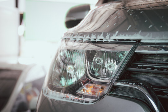 Modern White Car Covered By A Liquid Soap Foam While Washing. Headlight Of The Modern Car With A Soap Bobble And Form Close Up With Copyspace.