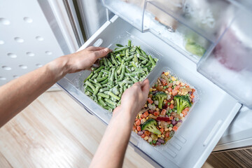 Hands are taking a container of frozen vegetables from the freezer of the fridge. Concept of...
