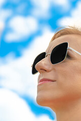 Blonde womans close-up profile face with black sunglasses and pink lipstick in blue sky with white clouds background with copy space