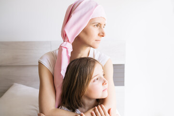 cancer mother with a pink headscarf hugging her blonde daughter looking at the window light
