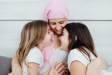 Cancer mother with a pink headscarf tenderly hugs her two daughters